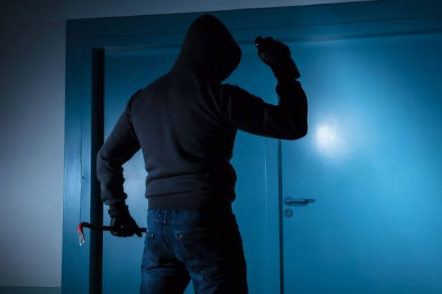 A hooded burglar holding a crowbar and a flashlight at a door in the dark.